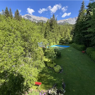 Ein schöner Garten mit üppigem Grün und bunten Blumen. Im Hintergrund sind Berge und ein blauer Pool zu sehen.