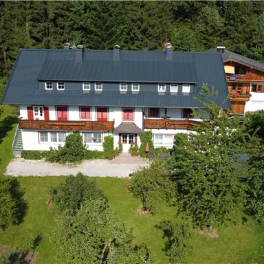 Ein großes, mehrstöckiges Haus mit einem grauen Dach umgeben von Bäumen und grünem Gras. Der Eingang hat eine einladende Terrasse und blühende Pflanzen.