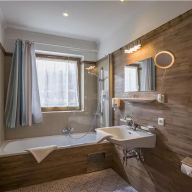 Ein modernes Badezimmer mit einer Badewanne und einem Waschbecken. Die Wände sind in Holzoptik gehalten, und es gibt viel natürliches Licht durch das Fenster.