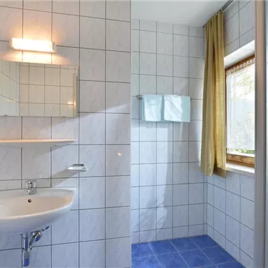 Ein modernes Badezimmer mit weißen Fliesen und einem blauen Bodenbelag. Es gibt ein Waschbecken, einen Spiegel und eine Duschkabine mit Fenstern, die natürliches Licht hereinlassen.