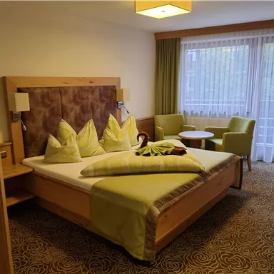 A modern hotel room with a large bed and green accents. Two cozy armchairs and a small table complement the furnishings.