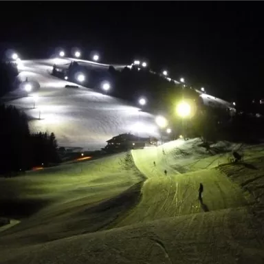 A illuminated ski slope at night with several skiers. The slope is surrounded by trees and the lights create a festive atmosphere.