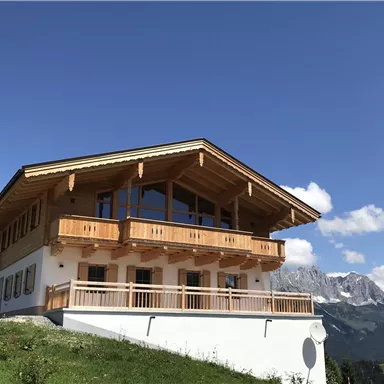A modern chalet in the mountains with wooden cladding and large windows. In the background, majestic mountains and a clear sky can be seen.