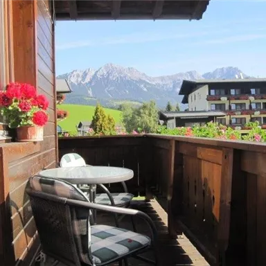 Ein gemütlicher Balkon mit einem Tisch und Stühlen, umgeben von bunten Blumen. Im Hintergrund sind die majestätischen Berge sichtbar.