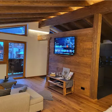 Ein modernes Wohnzimmer mit Holzverkleidung und großen Fenstern. Ein Fernseher hängt an der Wand, und die Möbel sind gemütlich und einladend.