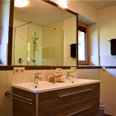 A modern bathroom with a wooden cabinet and a large mirror. The shower is separated by a glass wall, and two black towels are hanging on the wall.