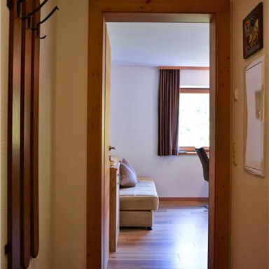 A bright hallway with wood paneling and a view of a cozy room. In the corner, there are skis, and there are large windows with curtains.