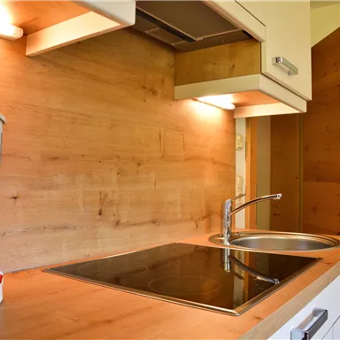 A modern kitchen with wooden surfaces and a cooktop. In the background, a coffee machine and a door are visible.