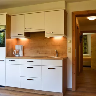 Modern kitchen with white cabinets and wooden details. In the background, access to another room is visible.