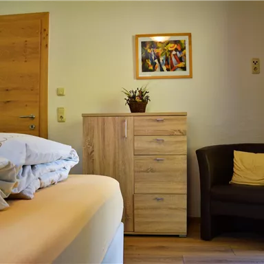 A cozy room with a bed, a dresser, and a chair. A colorful painting hangs on the wall.