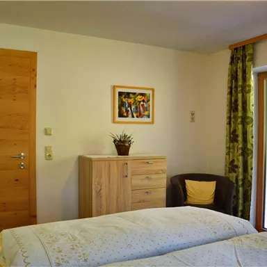 A cozy bedroom with two beds and a wooden wardrobe. The room has a balcony with a view of the outdoors and a beautiful piece of artwork on the wall.