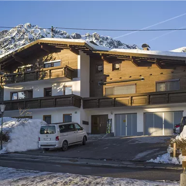 A modern wooden house in a snow-covered landscape with mountains in the background. There is a parking lot and clear blue skies.