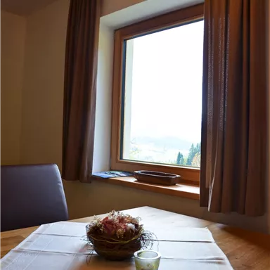 A dining area with a wooden table and a decorative dish in a nest. In the background, there is a window with a view of the landscape.
