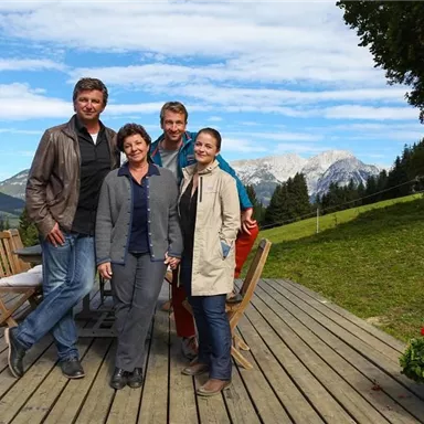 A group of four people stands on a terrace overlooking a picturesque landscape. In the background, mountains and a clear sky can be seen.