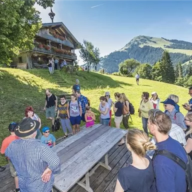 Eine Gruppe von Menschen versammelt sich im Freien vor einer Almhütte. Im Hintergrund sind grüne Hügel und eine klare blaue Himmelskulisse zu sehen.