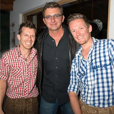 Drei Männer posieren zusammen vor einer Wand. Sie tragen traditionelle Trachten in karierten Mustern.