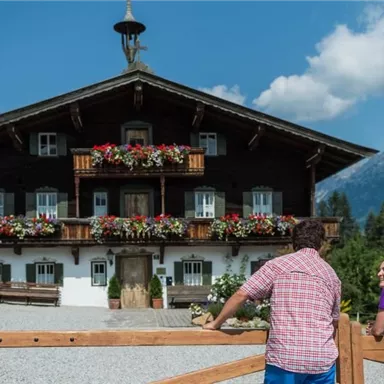 A traditional chalet with flowers on the balconies in front of a picturesque mountain landscape. Two people are standing by the fence and enjoying the view.