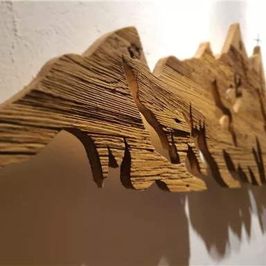 An artistic wooden relief depicting various mountain shapes. The structure and grain of the wood are clearly visible.
