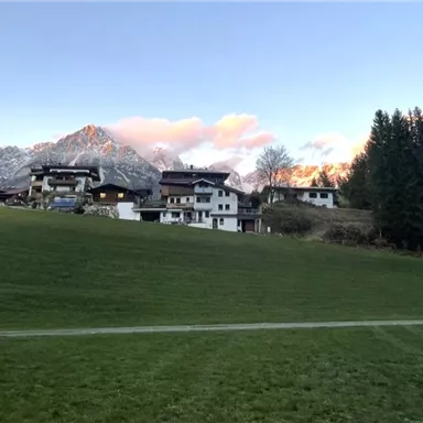 A picturesque landscape with houses in the foreground and mountains in the background. The sky is clear and the clouds are turning reddish.