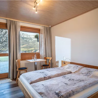 A cozy bedroom with a double bed and wooden elements. In daylight, a table with two chairs is visible, and large windows offer a view outside.
