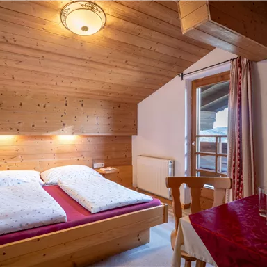 A cozy bedroom with wooden paneling and two beds. Through the window, one looks outside, and a table with chairs is also visible.