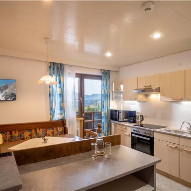 A modern kitchen with a cozy seating area and windows that lead to a balcony. Bright colors and a pleasant atmosphere create an inviting living environment.