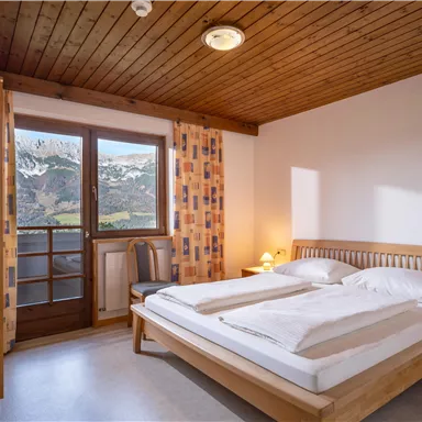 A bright bedroom with a large bed and wooden furniture. Windows provide a view of the mountains and nature.