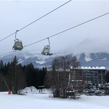 Eine Skiliftanlage mit Sesseln in einer schneebedeckten Landschaft. Im Hintergrund sind Berge und eine Wolkendecke zu sehen.