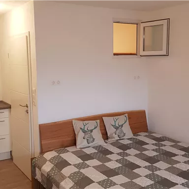 A cozy room with a large bed and a small kitchenette. The walls are bright and the window lets in natural light.