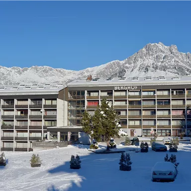 A modern building in the snow with high mountains in the background. The surroundings are wintry and clear with a blue sky.