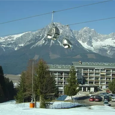 A picturesque mountain landscape with snow-covered peaks and a hotel in the foreground. A cable car floats above the scene.