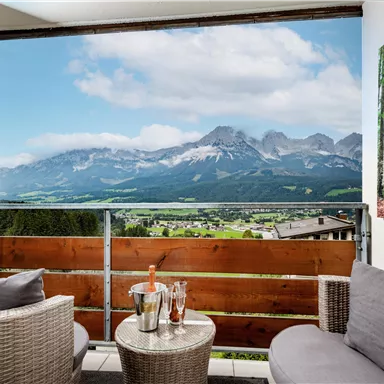 Ein gemütlicher Balkon mit zwei Sesseln und einem Tisch. Im Hintergrund erstrecken sich majestätische Berge unter einem teilweise bewölkten Himmel.