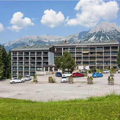 Ein modernes Gebäude mit mehreren Etagen in einer bergigen Landschaft. Im Vordergrund gibt es Parkplätze und viel Grünfläche.