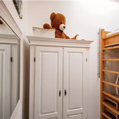 Ein gemütlicher Raum mit einem weißen Schrank und einem großen Teddy auf der oberen Ablage. Neben dem Schrank steht ein Holzregal.