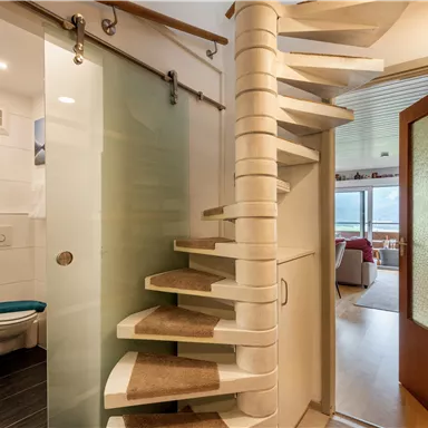 Eine moderne Treppe führt zu einem oberen Raum. Im Vordergrund sieht man einen hellen Badezimmerbereich mit einer Glaswand und einem Toilettenraum.