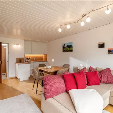 Ein modernes Wohnzimmer mit einem großen Sofa und bunten Kissen. Es gibt einen Essbereich und dekorative Bilder an der Wand.