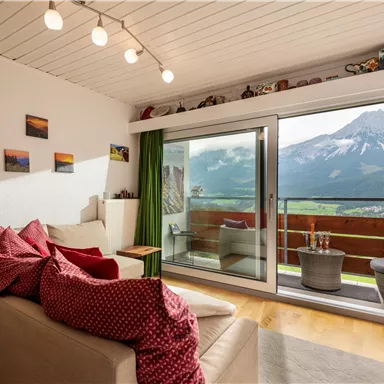 Ein gemütliches Wohnzimmer mit einer großen Fensterfront, die einen wunderschönen Blick auf die Berge bietet. Die Einrichtung ist modern und einladend, mit roten Kissen auf dem Sofa.