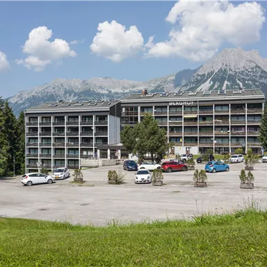 A modern building amidst mountains and green meadows. The parking lot in front of the building is well attended.