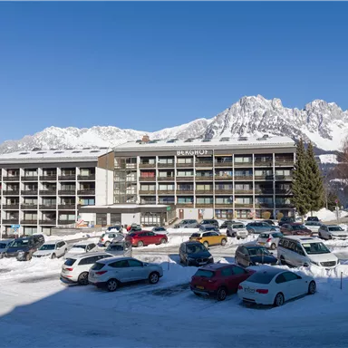 A modern hotel building in a snow-covered landscape. In the background, majestic mountains and a clear blue sky can be seen.