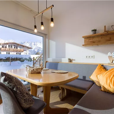 A cozy dining area with a large wooden table and comfortable benches. Through the window, you can see snow-covered mountains and a beautiful view.