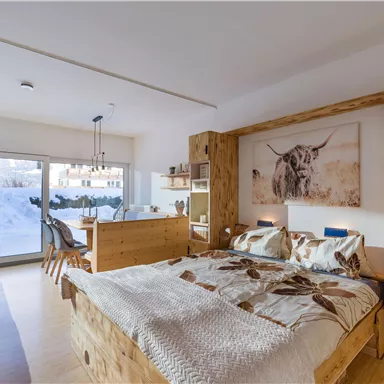 A modern bedroom with a large wooden bed and a cozy seating area. Large windows offer a view of a snow-covered outdoor area.