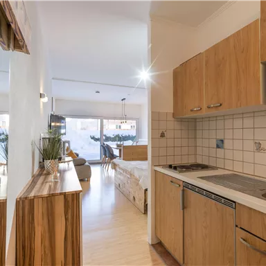 A modern kitchen unit with wooden details and white wall cladding. In the background, the living area is visible, filled with daylight and a cozy atmosphere.