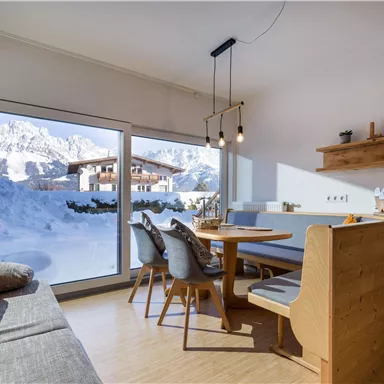 A modern living room with large windows and a view of snow-covered mountains. The space is cozy furnished with wooden details and a dining table.