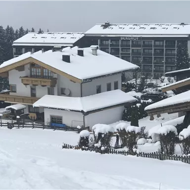 A snow-covered landscape with traditional mountain houses and a modern building in the background. The scene conveys a serene winter atmosphere.