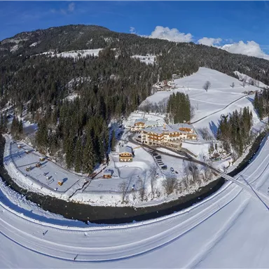 A picturesque winter landscape with snow-covered hills and a tranquil river. In the foreground, a charming accommodation is seen amidst nature.