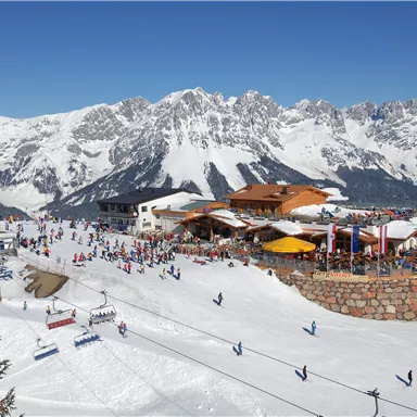 Eine winterliche Skilandschaft mit schneebedeckten Bergen und einer belebten Skihütte. Viele Menschen genießen den Schnee und die Sonne.