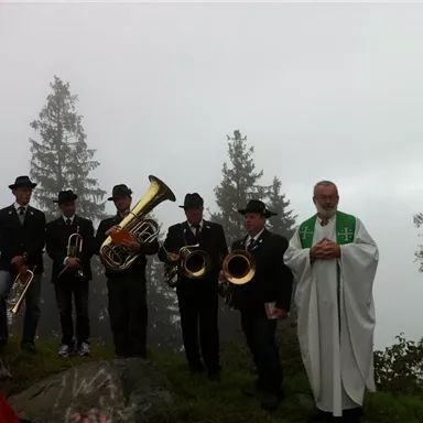 Eine Gruppe von Musikern steht auf einem bewaldeten Hügel inmitten von Nebel. Ein Priester steht vor ihnen und hält eine Zeremonie ab.