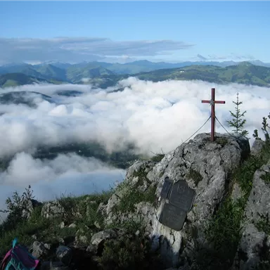 A view of a breathtaking mountain landscape with mist and clouds. In the foreground, there is a cross on a rock.