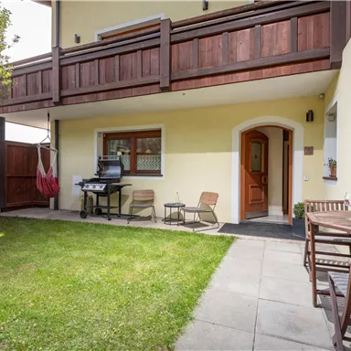 Ein gemütlicher Außenbereich mit einem kleinen Rasen, einem Grill und Holzstühlen. Die Umgebung ist von einem Holzzaun umgeben und es gibt einen hängenden Stuhl.