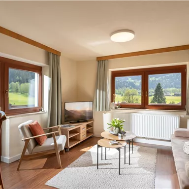 Ein gemütliches Wohnzimmer mit großen Fenstern und Blick auf die Landschaft. Die Einrichtung ist modern und einladend, mit einem Sofa und einem kleinen Tisch.